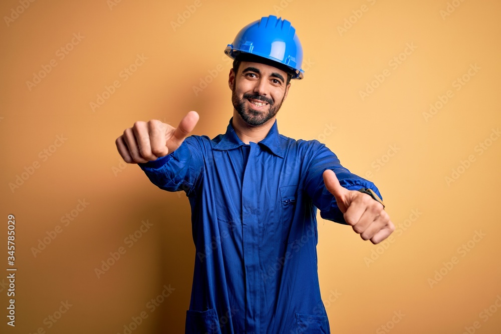 Mechanic man with beard wearing blue uniform and safety helmet over yellow background approving doing positive gesture with hand, thumbs up smiling and happy for success. Winner gesture.