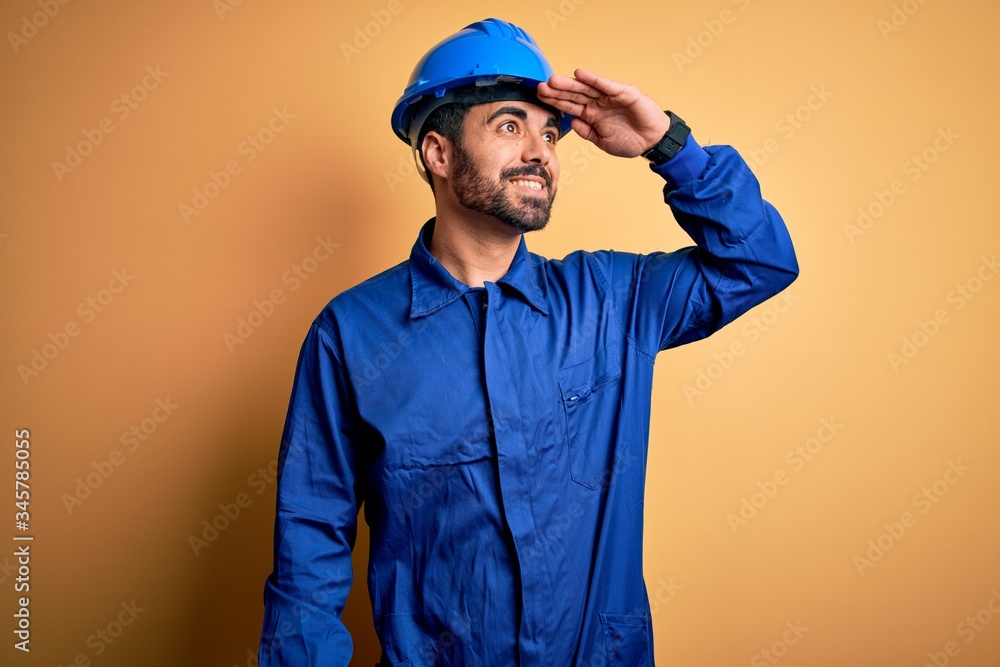 Mechanic man with beard wearing blue uniform and safety helmet over yellow background very happy and smiling looking far away with hand over head. Searching concept.