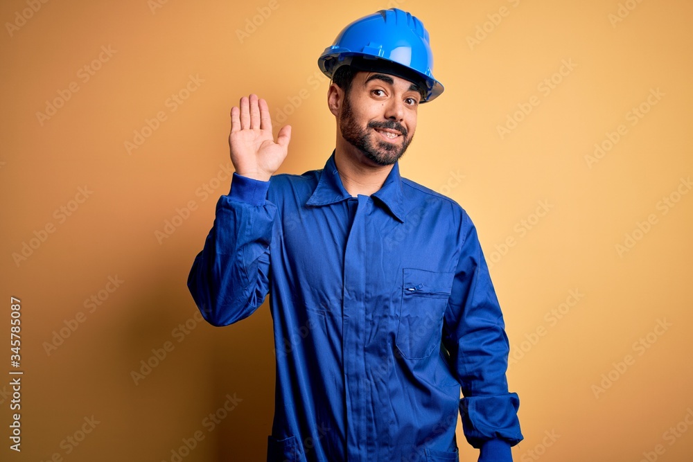 Mechanic man with beard wearing blue uniform and safety helmet over yellow background Waiving saying hello happy and smiling, friendly welcome gesture