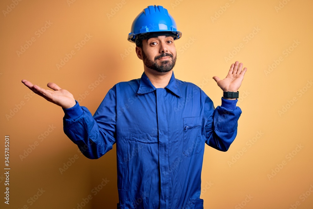 Mechanic man with beard wearing blue uniform and safety helmet over yellow background clueless and confused expression with arms and hands raised. Doubt concept.