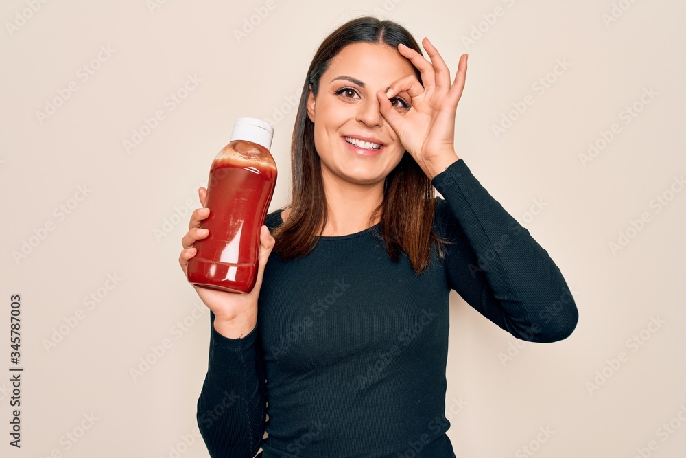 Young beautiful brunette woman holding bottle with ketchup sauce over white background smiling happy doing ok sign with hand on eye looking through fingers Young beautiful brunette woman holding bottle with ketchup sauce over white background smiling happy doing ok sign with hand on eye looking through fingers