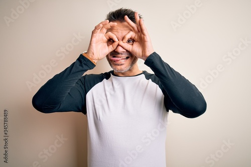 Young handsome man wearing casual t-shirt standing over isolated white background doing ok gesture like binoculars sticking tongue out, eyes looking through fingers. Crazy expression.