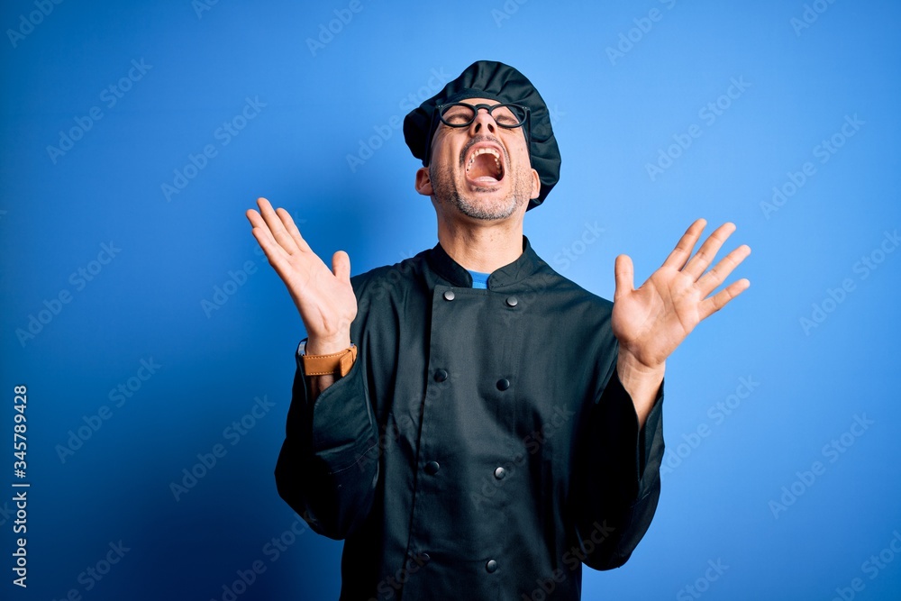 Young handsome chef man wearing cooker uniform and hat over isolated blue background celebrating mad and crazy for success with arms raised and closed eyes screaming excited. Winner concept Young handsome chef man wearing cooker uniform and hat over isolated blue background celebrating mad and crazy for success with arms raised and closed eyes screaming excited. Winner concept