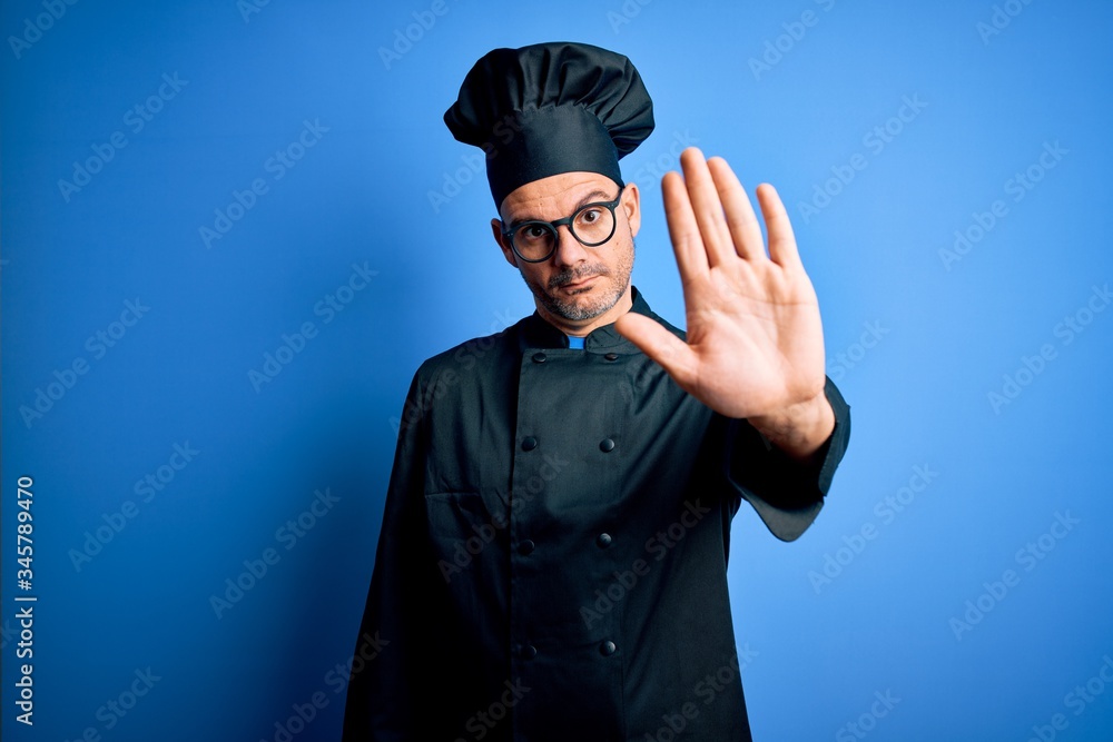 Young handsome chef man wearing cooker uniform and hat over isolated blue background doing stop sing with palm of the hand. Warning expression with negative and serious gesture on the face. Young handsome chef man wearing cooker uniform and hat over isolated blue background doing stop sing with palm of the hand. Warning expression with negative and serious gesture on the face.
