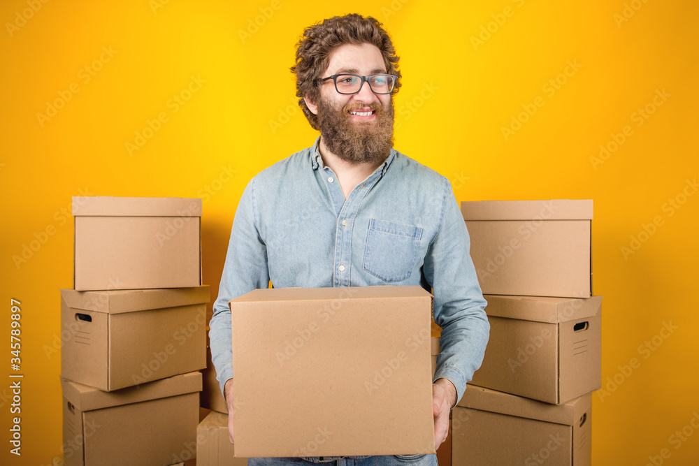 Delivery man holding a cardboard box in his hands standing among many boxes with parcels on a yellow background Delivery man holding a cardboard box in his hands standing among many boxes with parcels on a yellow background