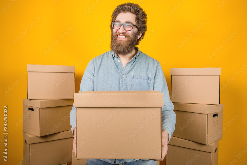 Bearded male delivery man holding a corton box in his hands, standing among parcels on a yellow background. Goods delivery Bearded male delivery man holding a corton box in his hands, standing among parcels on a yellow background. Goods delivery