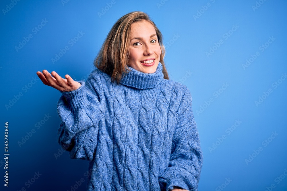 Young beautiful blonde woman wearing casual turtleneck sweater over blue background smiling cheerful with open arms as friendly welcome, positive and confident greetings