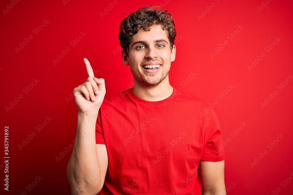 Young blond handsome man with curly hair wearing casual t-shirt over red background pointing finger up with successful idea. Exited and happy. Number one. Young blond handsome man with curly hair wearing casual t-shirt over red background pointing finger up with successful idea. Exited and happy. Number one.