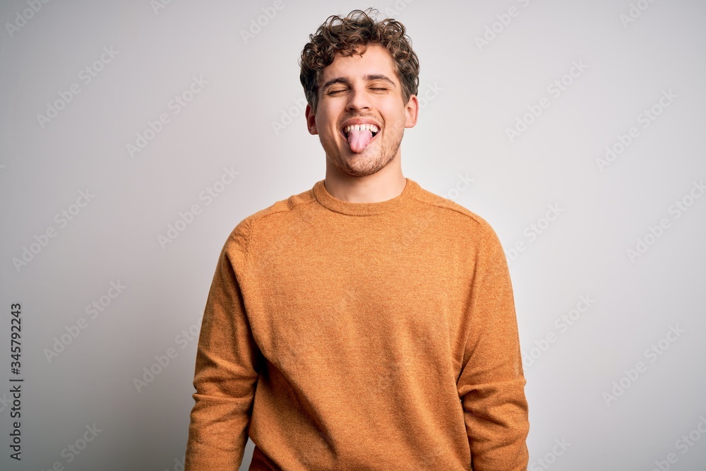 Young blond handsome man with curly hair wearing casual sweater over white background sticking tongue out happy with funny expression. Emotion concept. Young blond handsome man with curly hair wearing casual sweater over white background sticking tongue out happy with funny expression. Emotion concept.