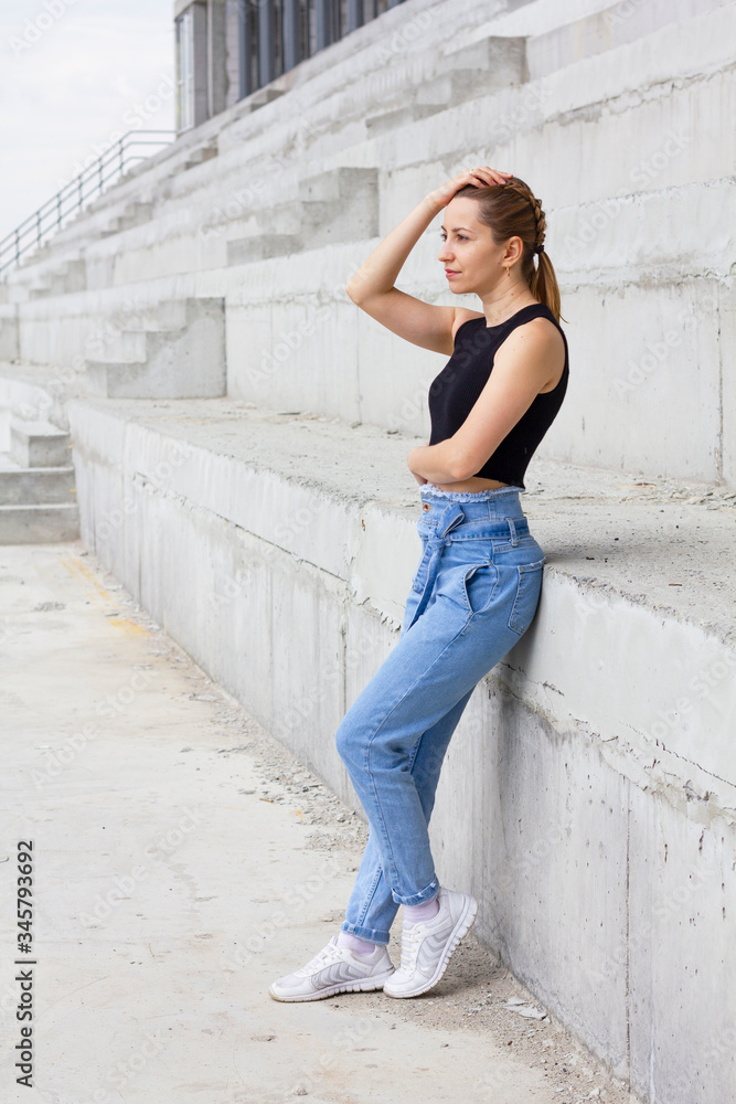 Gorgeous fashion sexy female dressed in black cropp top and blue jeans standing outdoor, smiling and enjoying sunny day. Sports arena in the background. Girl posing at sunny day. Gorgeous fashion sexy female dressed in black cropp top and blue jeans standing outdoor, smiling and enjoying sunny day. Sports arena in the background. Girl posing at sunny day.