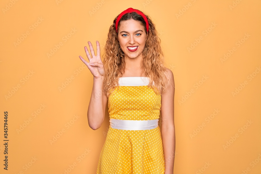 Obraz premium Beautiful blonde pin-up woman with blue eyes wearing diadem standing over yellow background showing and pointing up with fingers number four while smiling confident and happy.
