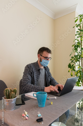 Side view of young european man wearing a face-mask working from home due to the corona-virus restrictions using the laptop