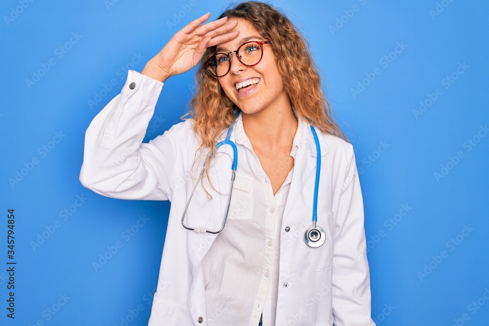 Young beautiful blonde doctor woman with blue eyes wearing coat and stethoscope very happy and smiling looking far away with hand over head. Searching concept. Young beautiful blonde doctor woman with blue eyes wearing coat and stethoscope very happy and smiling looking far away with hand over head. Searching concept.