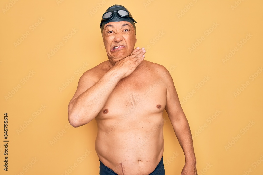 Middle age senior grey-haired swimmer man wearing swimsuit, cap and goggles cutting throat with hand as knife, threaten aggression with furious violence