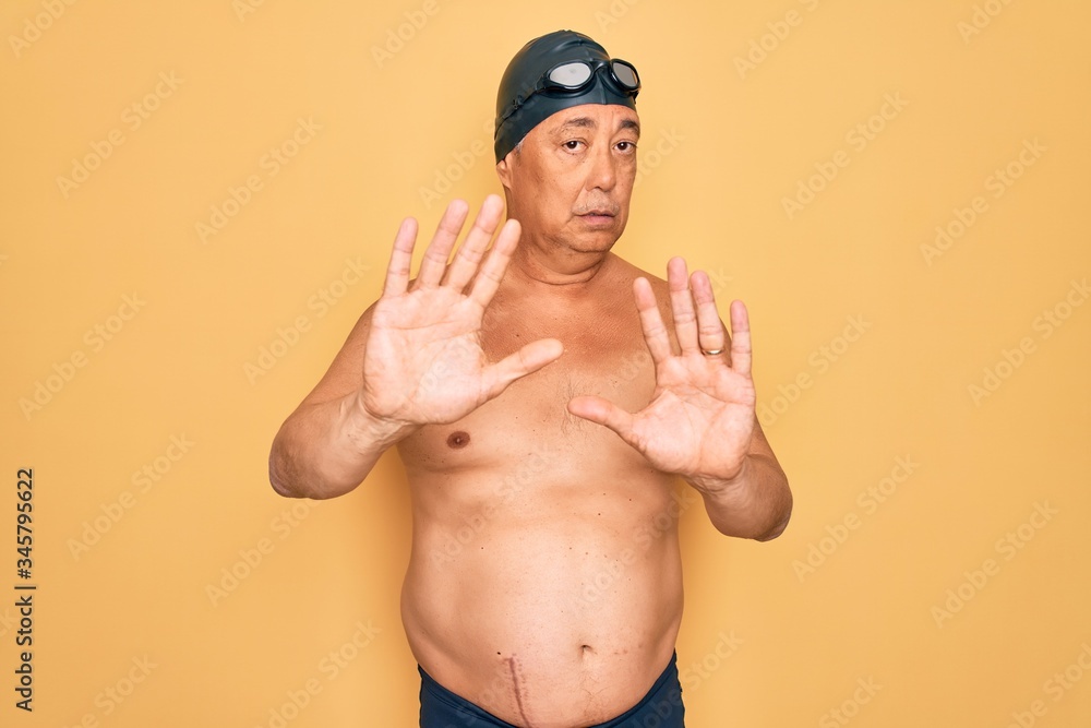 Middle age senior grey-haired swimmer man wearing swimsuit, cap and goggles Moving away hands palms showing refusal and denial with afraid and disgusting expression. Stop and forbidden.