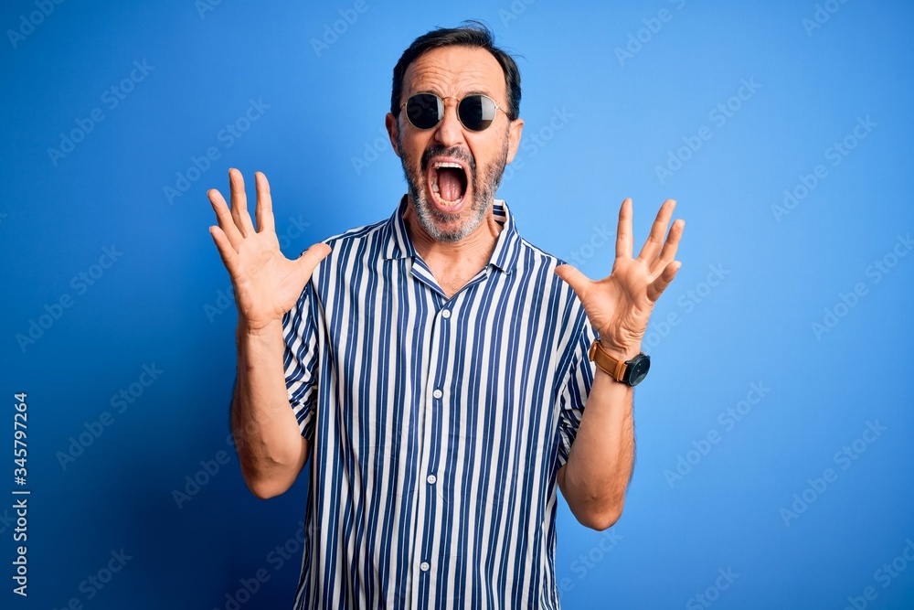 Middle age hoary man wearing striped shirt and sunglasses over isolated blue background crazy and mad shouting and yelling with aggressive expression and arms raised. Frustration concept. Middle age hoary man wearing striped shirt and sunglasses over isolated blue background crazy and mad shouting and yelling with aggressive expression and arms raised. Frustration concept.