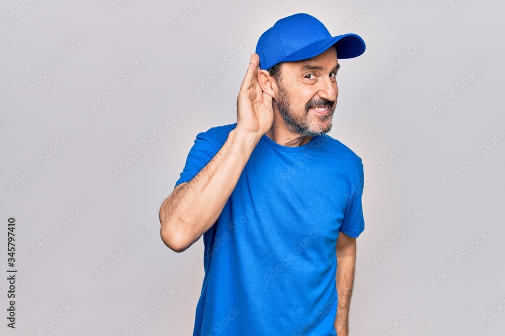 Middle age handsome deliveryman wearing cap standing over isolated white background smiling with hand over ear listening and hearing to rumor or gossip. Deafness concept. Middle age handsome deliveryman wearing cap standing over isolated white background smiling with hand over ear listening and hearing to rumor or gossip. Deafness concept.