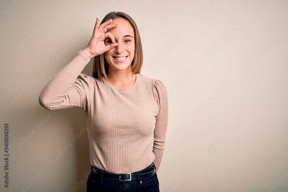 Young beautiful woman wearing casual sweater standing over isolated white background doing ok gesture with hand smiling, eye looking through fingers with happy face. Young beautiful woman wearing casual sweater standing over isolated white background doing ok gesture with hand smiling, eye looking through fingers with happy face.