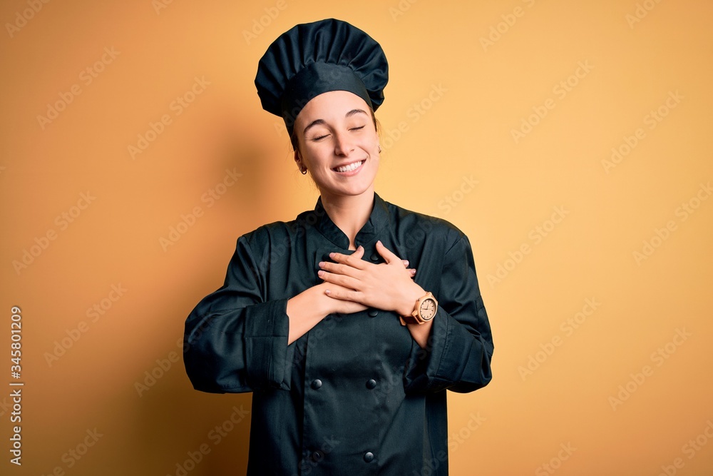 Young beautiful chef woman wearing cooker uniform and hat standing over yellow background smiling with hands on chest with closed eyes and grateful gesture on face. Health concept. Young beautiful chef woman wearing cooker uniform and hat standing over yellow background smiling with hands on chest with closed eyes and grateful gesture on face. Health concept.