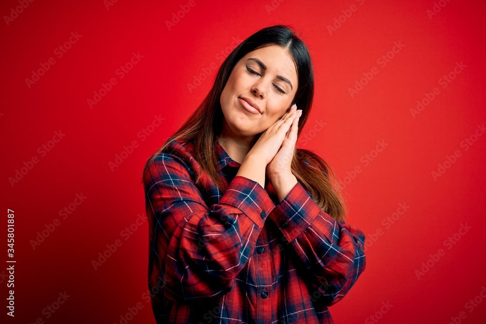 Young beautiful woman wearing casual shirt over red background sleeping tired dreaming and posing with hands together while smiling with closed eyes. Young beautiful woman wearing casual shirt over red background sleeping tired dreaming and posing with hands together while smiling with closed eyes.