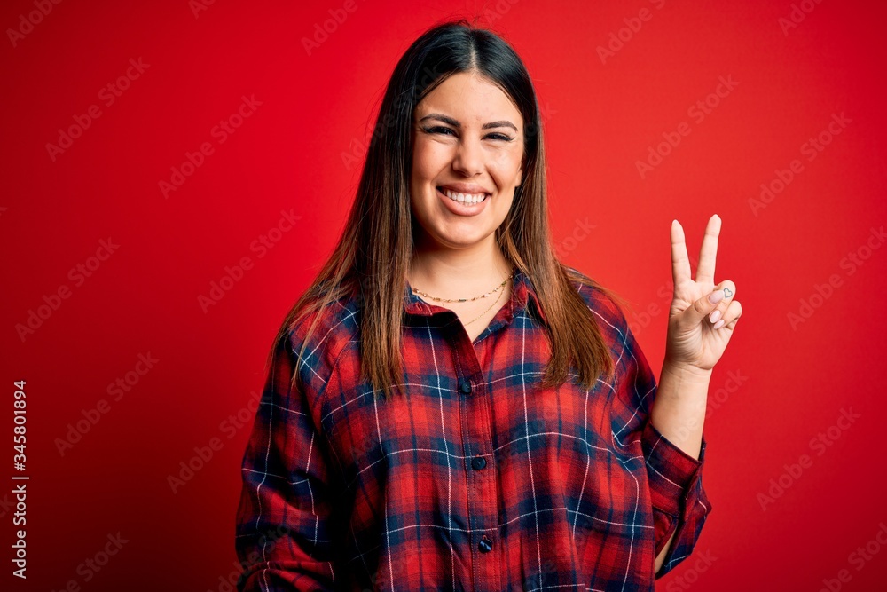 Young beautiful woman wearing casual shirt over red background smiling with happy face winking at the camera doing victory sign. Number two. Young beautiful woman wearing casual shirt over red background smiling with happy face winking at the camera doing victory sign. Number two.