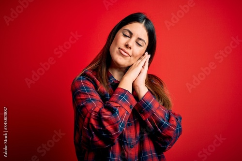 Young beautiful woman wearing casual shirt over red background sleeping tired dreaming and posing with hands together while smiling with closed eyes. © Krakenimages.com