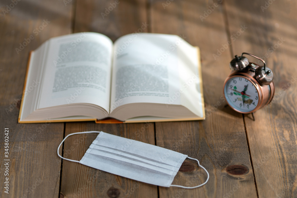 A hardback book opened for reading and a desk clock next to it. Clock book and mask on wooden background A hardback book opened for reading and a desk clock next to it. Clock book and mask on wooden background