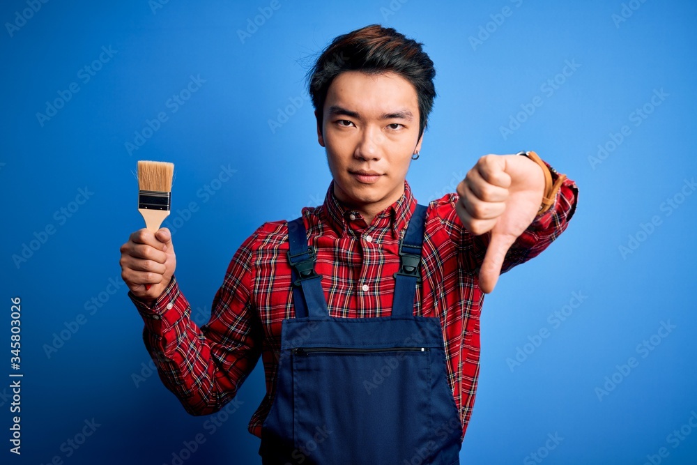 Young handsome chinese painter man wearing apron painting using brush with angry face, negative sign showing dislike with thumbs down, rejection concept Young handsome chinese painter man wearing apron painting using brush with angry face, negative sign showing dislike with thumbs down, rejection concept