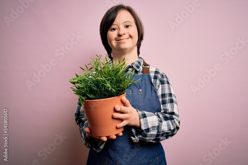 Young down syndrome gardener woman wearing worker apron holding green plant pot with a happy face standing and smiling with a confident smile showing teeth