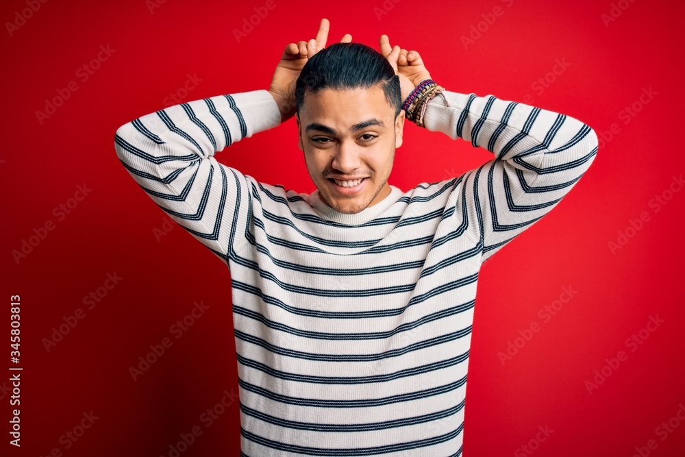 Young brazilian man wearing casual striped t-shirt standing over isolated red background Posing funny and crazy with fingers on head as bunny ears, smiling cheerful