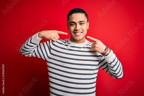 Young brazilian man wearing casual striped t-shirt standing over isolated red background smiling cheerful showing and pointing with fingers teeth and mouth. Dental health concept.
