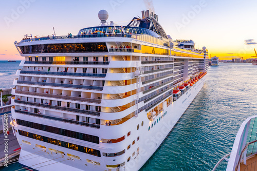 Back/ Stern and starboard side of Cruise Ship docked/ anchored/ moored in Port with other ships. People on decks. Amazing sunset sky in background.