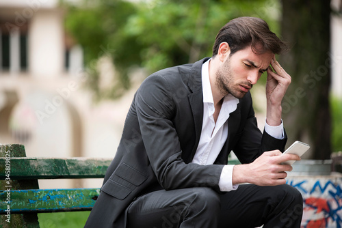 Attractive young businessman sitting on a bench in the park, receiving some disturbing news on his telephone