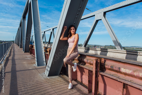 Pretty young woman posing on the old rusty transport bridge over the river during sunset. © Smile