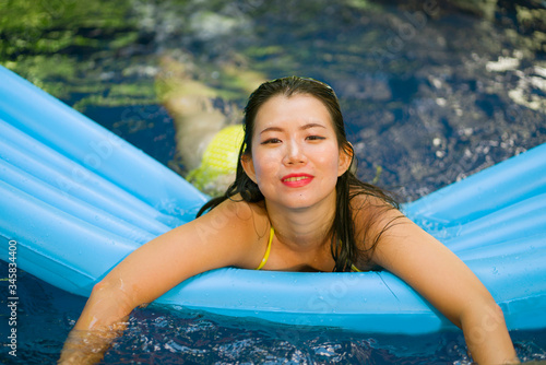  young beautiful and happy Asian Korean woman in bikini enjoying Summer holiday at swimming pool playful having fun on airbed at villa or luxury tropical resort