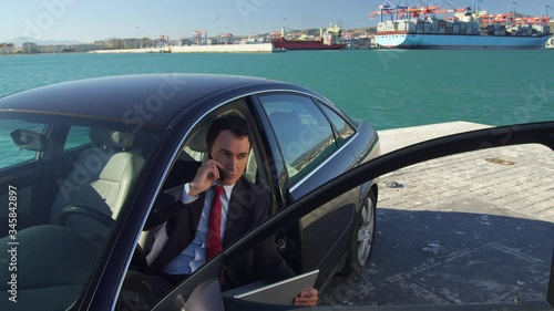 WS Businessman sitting in car on quayside in port, using laptop and phone