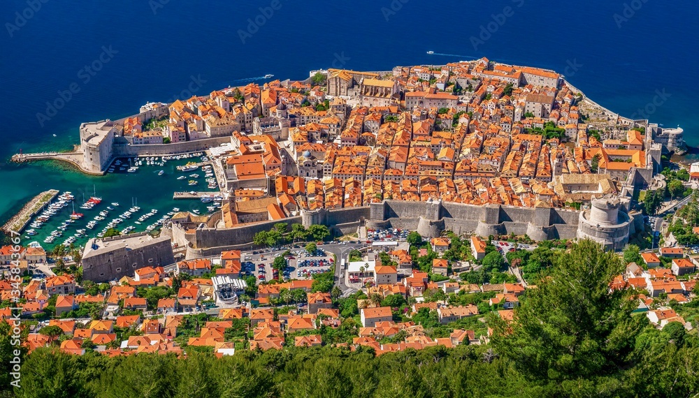 A high angle view of the entire Old Town of Dubrovnik, Croatia, with its terracotta rooftops, surrounded by stone walls and a small boat harbor open to the Adriatic Sea.