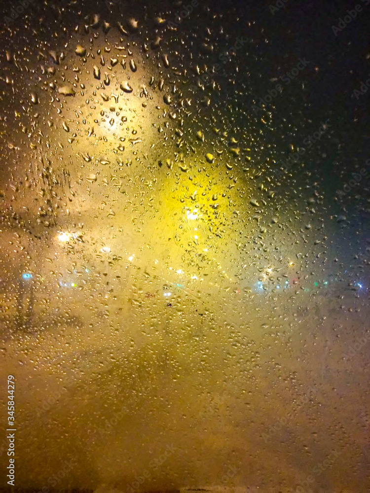 Drops of rain on window at night. Rainy and night city behind glass. Tears of the sky flow down transparent wall. Bead drops hang in light of lanterns and Windows. Drops of rain on window at night. Rainy and night city behind glass. Tears of the sky flow down transparent wall. Bead drops hang in light of lanterns and Windows.