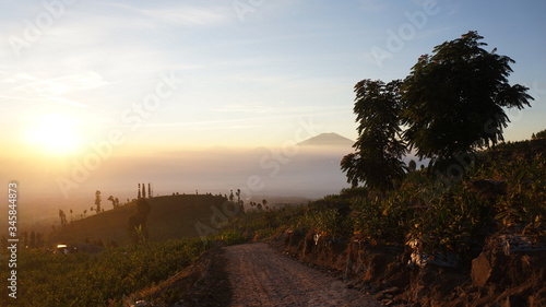 view of a road on the slopes of Mount Sindoro at sunrise with the background of Mount Merapi Merbabu covered in white clouds. Photo taken in July 2019