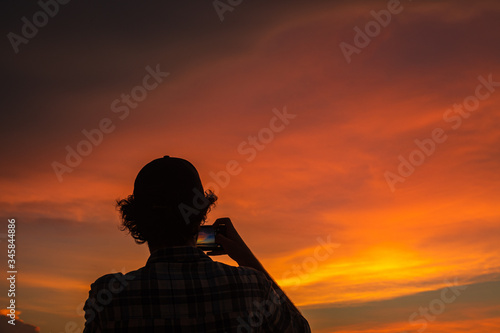 silhouette of a young man wearing baseball cap taking photo of beautiful sunset.