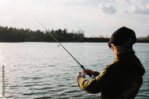 Fisherman with rod, spinning reel on the river bank. Sunset. Fishing for pike, perch, carp. Woman catching a fish, pulling rod while fishing. Girl fishing from beach lake or pond with text space.