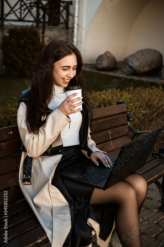 Portrait of a glamorous young woman holding on her knees portable laptop computer while sitting on a wooden bench, stylish female drinking coffee while relaxing after work on net-book during free time