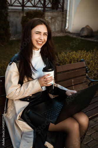 close portrait of a girl with a laptop sitting on a bench with a cup of coffee. Student with laptop studying in the street.