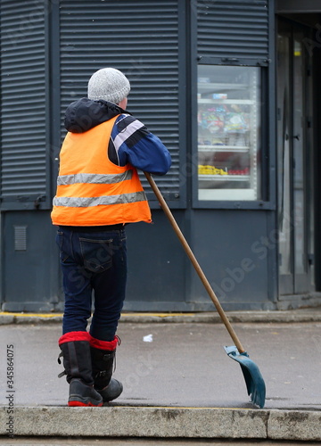 Janitor in an orange vest is leaning on a shovel