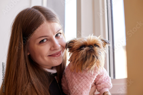 Girl with long hair in a black jacket and a small dog in pink clothes at the window indoors.