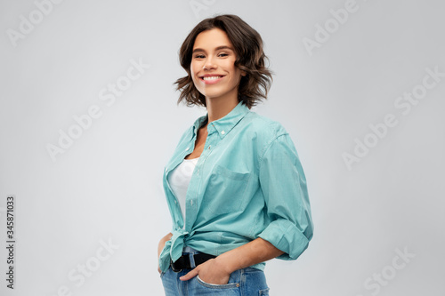 people concept - portrait of happy smiling young woman in turquoise shirt with hands in pockets over grey background