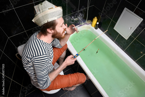 home isolation and quarantine concept. holiday at home, man catches fish in bath. newspaper cap with unreadable text
