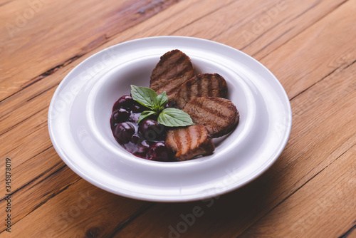 Grilled meat pieces with berry sauce served on a white plate over dark rustic wooden background. Italian cuisine.