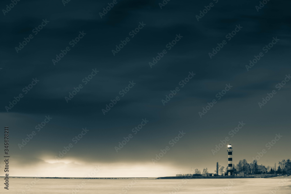 Sea waves of the sea at long exposure. Cloudy sky over the water. Striped lighthouse on the seashore. Toned black and white.