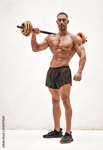 Shirtless adult male bodybuilder doing an exercise with a barbell in a bright studio